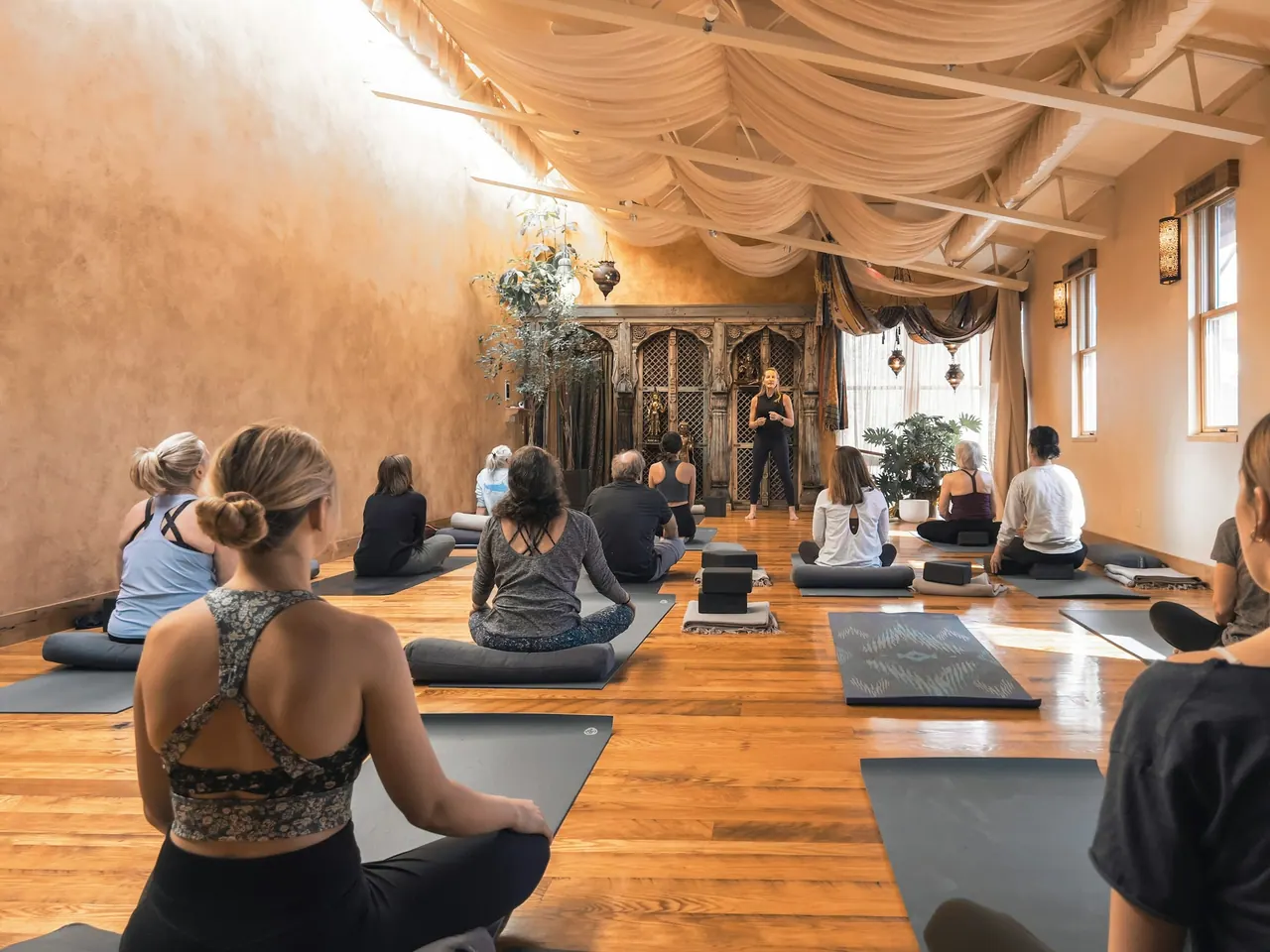 A guided class seated on mats in a warm hall with draped ceilings