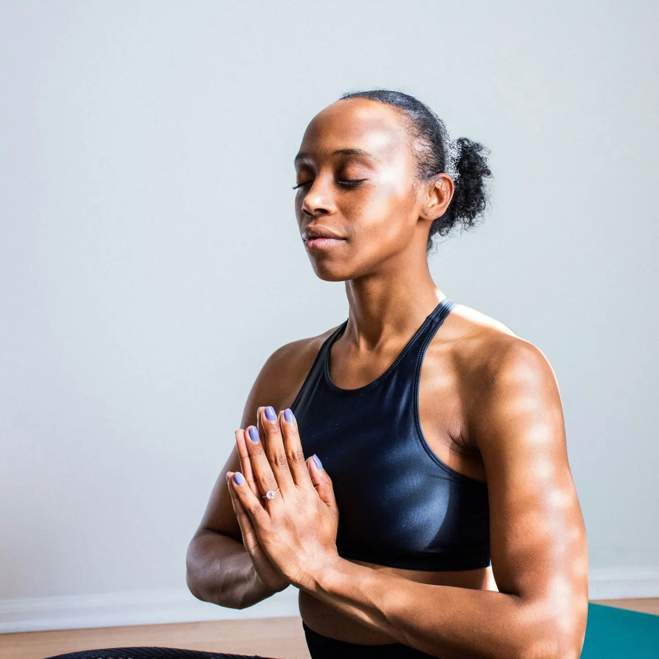 A woman sitting tall with her palms together and her eyes closed, in soft window light